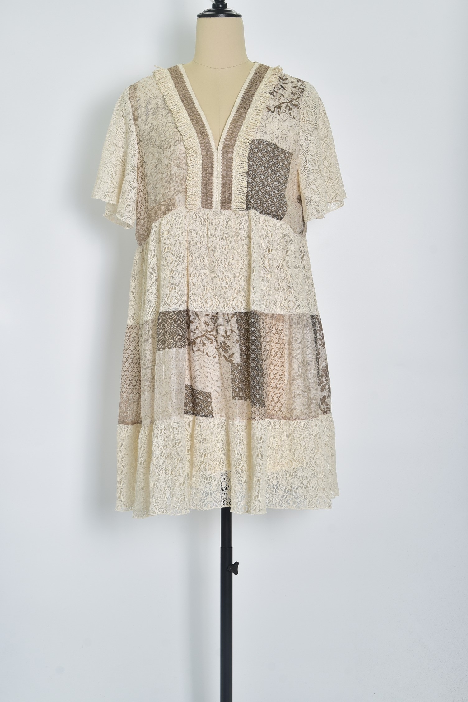 Réf. 41002 - Robe en dentelle patchwork, combinant différentes textures et motif dans des couleurs neutres. Décolleté plongeant en V et des manches courtes légèrement évasées, elle dégage un charme bohème et féminin. La robe s'évase en une jupe ample à volants, ce qui crée une silhouette gracieuse et aérienne. C'est une tenue parfaite pour les événements printaniers ou estivaux, alliant élégance et confort.